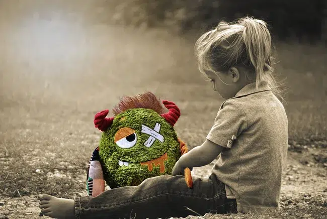 Une jeune enfant joue avec une peluche verte, abîmée et avec un œil en moins. Elle trouve plus de réconfort à s’exprimer à travers elle, projetant ainsi sa propre souffrance.